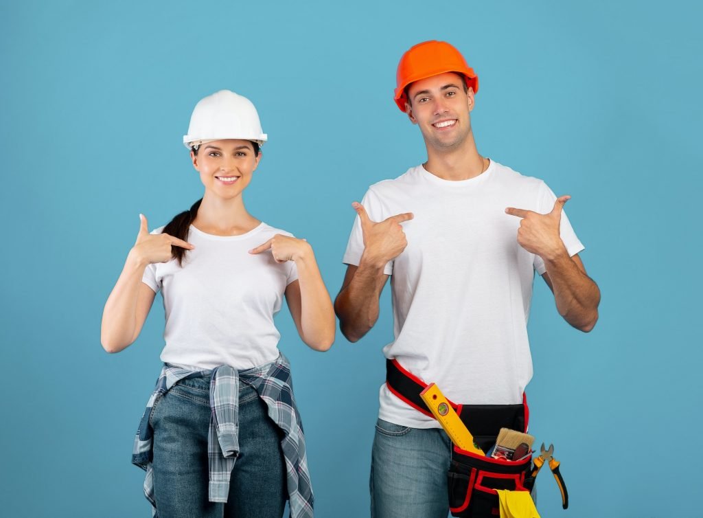 Choose Us For Repair. Two Happy Handyworkers In Hardhats Pointing At Themselves